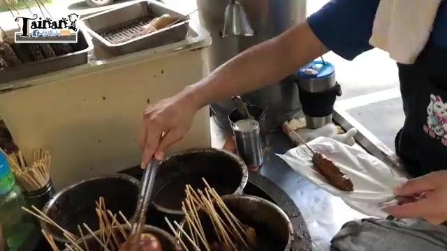 老闆手持竹籤將串好的黑輪放入醬料鍋中翻動沾醬，一旁堆疊著待處理的竹籤