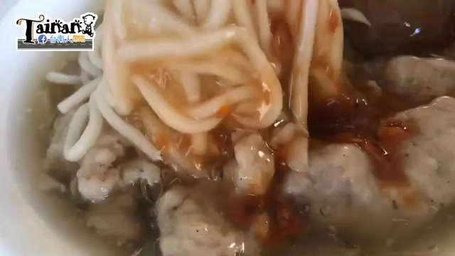 特寫麵條與肉羹在湯中,麵條呈現淡黃色澤,表面沾附紅色油亮醬汁與肉羹碎塊