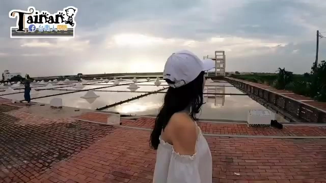 鹽田水面倒映天空雲層,遠處可見三角形鹽堆與瞭望台,遊客站在磚紅色步道上眺望