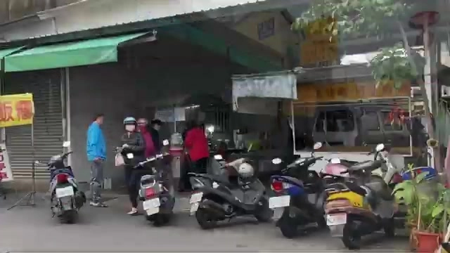 店家綠色遮陽篷下擺著攤位，摩托車停靠在店門口，早晨顧客正在排隊