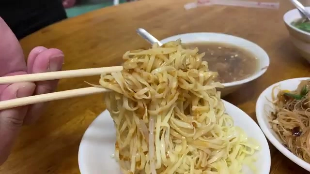 筷子夾起滿滿一把拌麵條，麵條上還帶著豆芽，一旁擺著湯碗與另一盤米粉