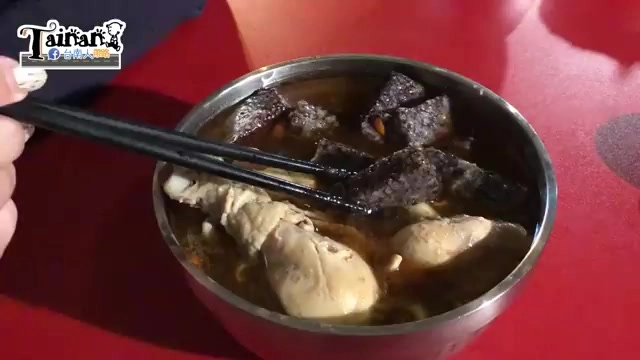 不鏽鋼碗盛著麻油雞湯，湯色深褐，可見雞腿肉塊與深色米血，筷子夾起一塊米血