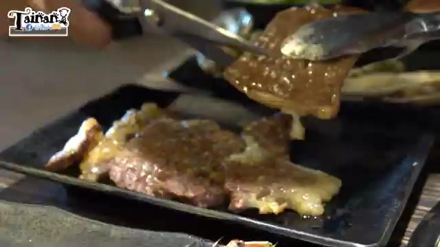 黑色餐盤盛著烤好的沙朗牛排，正被夾子夾起淋上醬汁，肉質呈現焦香色澤