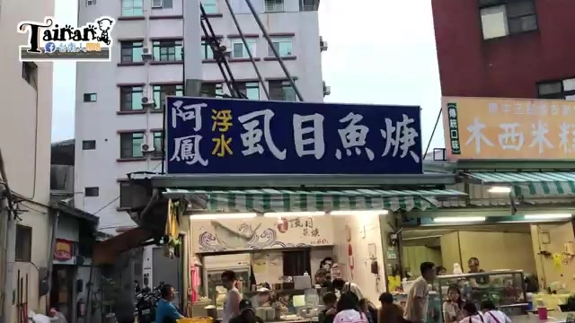 店家招牌，藍底白字寫著「阿鳳浮水虱目魚羹」，攤位前聚集排隊人潮，周邊還有其他店家招牌