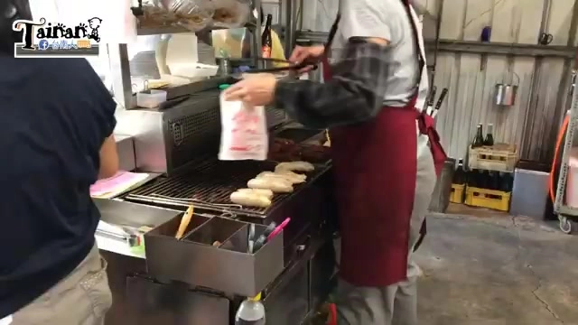 炭烤架上正在燒烤香腸，老闆穿著圍裙一手拿夾子翻烤，一手拿著裝滿食材的塑膠袋