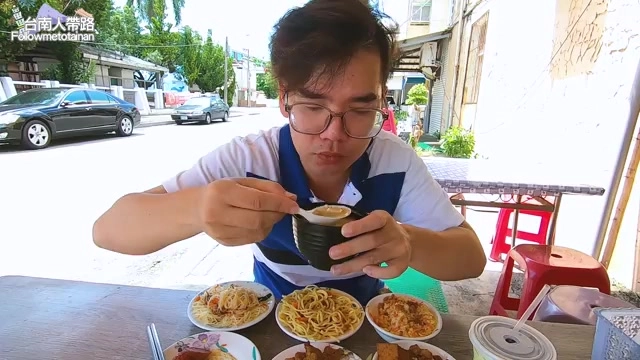 戴眼鏡的男性主持人坐在戶外座位，手捧黑色碗品嚐味噌湯，桌上擺著多碗米粉、麵條與滷味小菜
