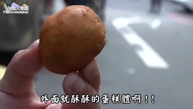 手持一顆金黃色圓形炸物，外皮酥脆帶油亮光澤，背景為街道人行道