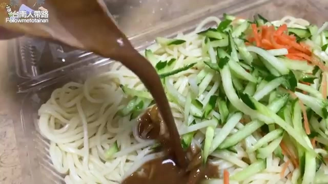 透明餐盒中盛著白色涼麵，正淋上深色麻醬，一旁堆疊著小黃瓜絲與紅蘿蔔絲
