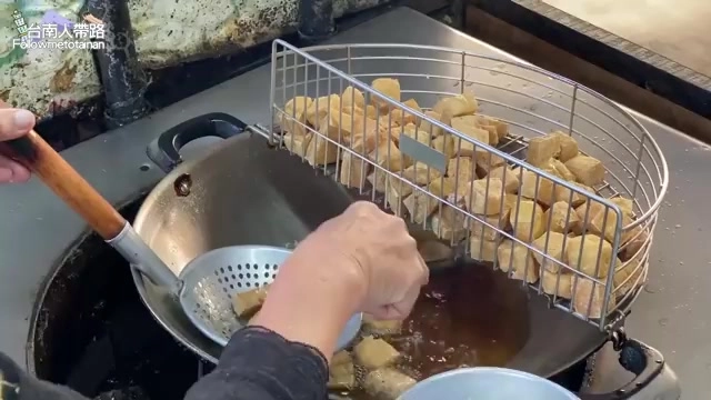 鐵籃中盛裝正在油炸的臭豆腐塊，老闆的手正在攪拌鍋中的油炸物