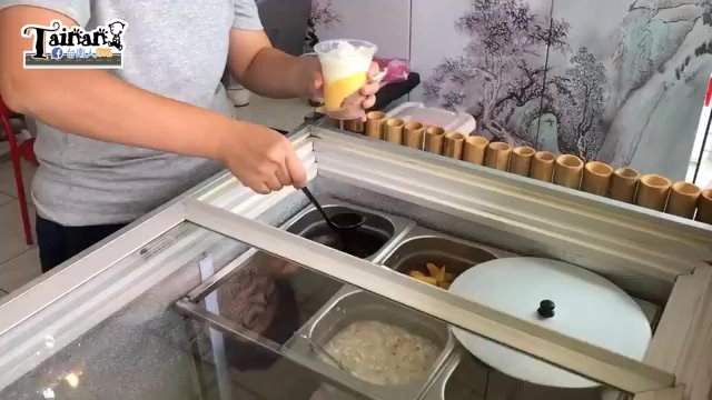 店員在冷藏櫃前製作飲品，手持透明杯裝著黃白分層飲料，冷藏格內盛裝黑糖水與配料