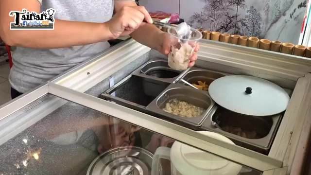 店員手持塑膠杯盛裝配料，冷藏格內分裝著紅豆、薏仁等多種配料，一旁放著白色圓形容器