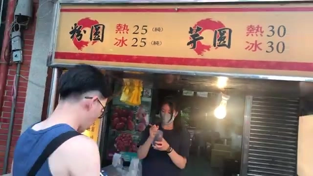 店面招牌列出粉圓與芋圓的冰熱價格，老闆在店內忙碌準備，貨架擺放紅色包裝商品