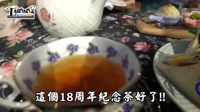 白色描花瓷茶杯裝著琥珀色紅茶，桌布印著玫瑰花紋，一旁擺著白色茶壺與其他茶點