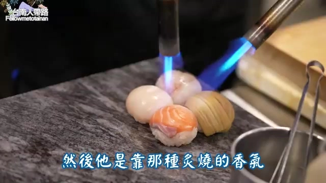 噴槍火焰正在炙燒握壽司表面，鮭魚與干貝配料清晰可見，冒出陣陣熱氣