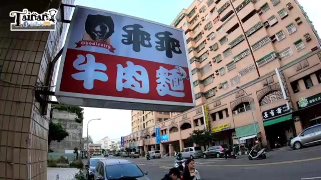紅底招牌上以粗黑字寫著「乖乖牛肉麵」，搭配可愛熊貓圖案，掛在街角建築外牆