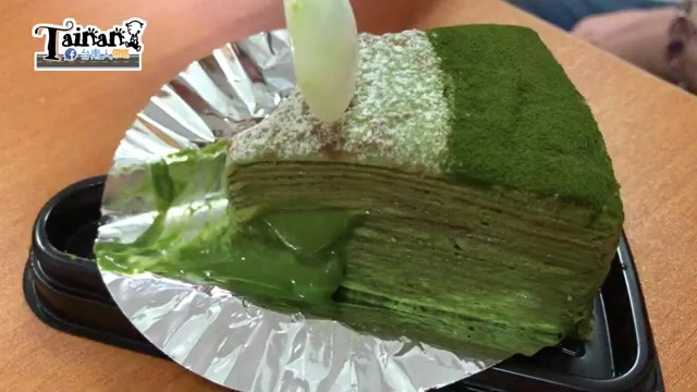 切開的抹茶千層蛋糕特寫，表面撒滿抹茶粉，切面可見層層薄餅與濃郁抹茶醬汁溢出盤外