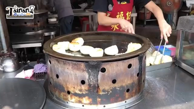 黑色圓形鐵鍋上正在煎製多顆水煎包,表皮呈現金黃色澤,老闆手持夾子在一旁翻動