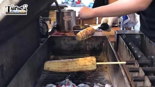 烤架上正在燒烤兩支玉米,店員手持夾具翻動,木炭火烤出誘人焦香