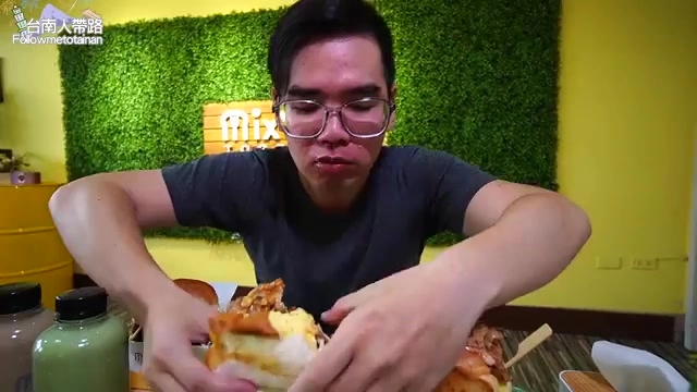 男性主持人雙手捧著厚實的吐司三明治，比自己臉還大，背景是餐廳的綠色植生牆