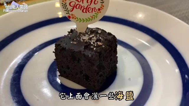 白色圓盤上擺著一塊方形巧克力布朗尼，插著印有店家logo的裝飾牌，表面撒著海鹽結晶