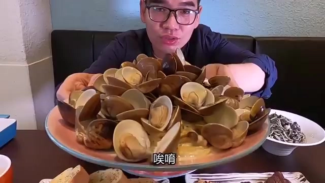 男性主持人坐在餐廳裡,面前擺著堆成小山的蛤蜊義大利麵
