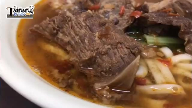 白色瓷碗盛著紅燒牛肉麵，湯色深褐帶紅辣椒點綴，牛肉塊與寬麵交疊