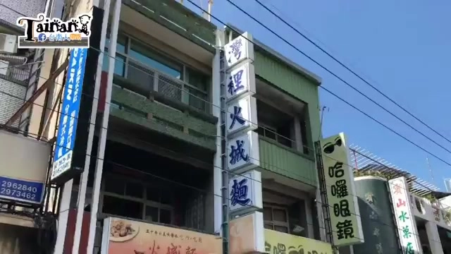 直立式店招牌，白底綠字寫著「灣裡火城麵」，一旁還有其他店家招牌與哈囉眼鏡的看板