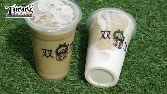 草地上放著兩杯綠豆沙飲品，透明杯身印有「双生」品牌圖樣，一杯是原味綠豆沙、一杯是綠豆沙牛奶