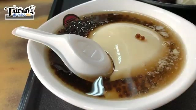 白色瓷碗盛著豆花,淋上深褐色糖水,可見黑色粉圓與白色豆花對比鮮明,一旁擺著白色湯匙