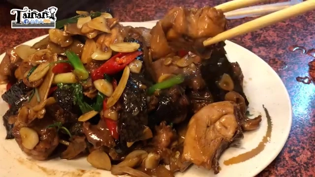 白色瓷盤盛著三杯雞，可見雞肉塊與深色米血搭配蒜片、辣椒與九層塔，醬色濃郁油亮，筷子夾起一塊雞肉
