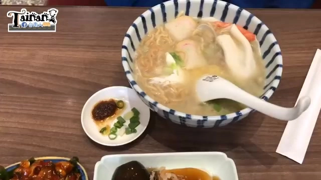 藍白條紋瓷碗盛著鍋燒意麵，可見蝦子、魚板、豆腐等配料，旁邊擺著兩碟小菜與沙茶蔥花沾料