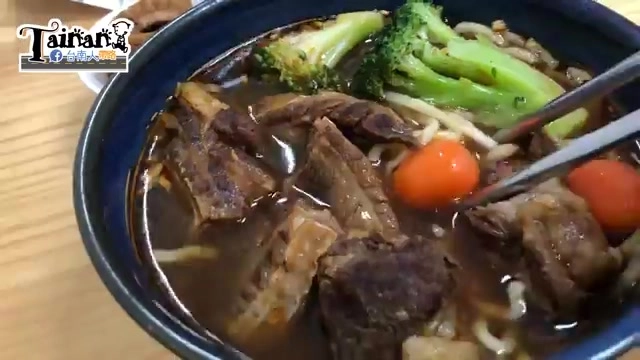 深藍色瓷碗盛著紅燒牛肉麵，可見大塊牛肉、綠花椰菜與小番茄，筷子夾起一塊肉