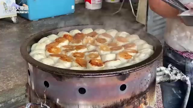 大鐵鍋裡整齊排列著正在半煎炸的水煎包，冒著蒸氣，老闆在一旁翻動鍋鏟