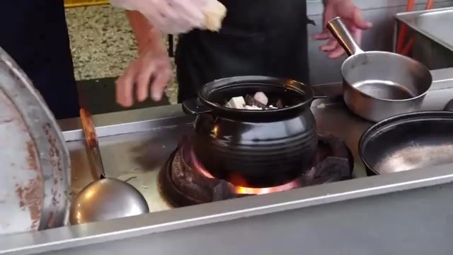 黑色陶鍋架在炭火爐上燉煮，師傅正在加入食材，爐下炭火明顯燃燒