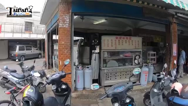 店面外觀，鐵板架上擺著菜單價目表，門口停放多台機車，瓦斯桶整齊排列在牆邊