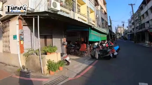 巷弄轉角的店面，綠色遮陽棚下擺著桌椅，機車停放在路邊，招牌不明顯但氛圍老派懷舊
