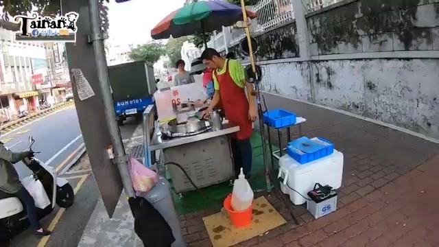 路邊攤販前撐著遮陽傘，老闆正在鐵板上製作烙餅，攤位旁擺放保溫箱與調味料