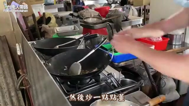 廚房裡老闆正在爐火前翻炒鍋中食材,兩口炒鍋並排使用,蒸氣裊裊