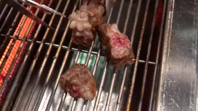 燒烤架上正在炙烤數塊牛肉,肉質可見粉紅色澤與白色油脂,架下炭火通紅